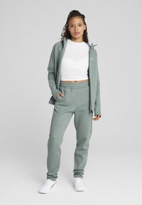Survêtement de sport vert clair avec une veste zippée et un pantalon à taille élastique. Top court blanc en dessous, assorti à des baskets blanches.