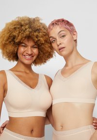 Dos mujeres diversas vestidas con sujetadores y ropa interior de encaje beige a juego, de pie juntas y sonriendo contra un fondo simple.
