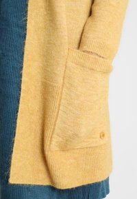 Cardigan in morbido giallo di lana con una texture a costine, dotato di una tasca laterale e dettagli con bottoni. Abbinato a un tessuto in teal a costine sotto.