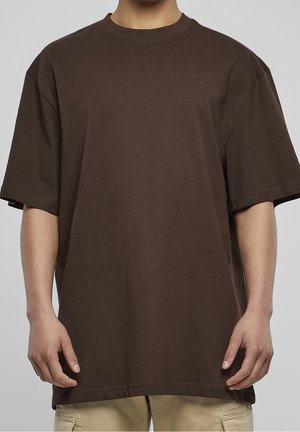 Personne portant un t-shirt ample marron foncé à manches courtes et un pantalon beige sur un fond gris uni.