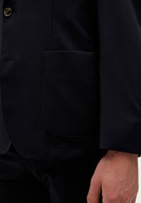 Blazer navy con cierre de un solo botón y un gran bolsillo redondeado. Tejido suave con una textura sutil; también muestra parte de una mano.