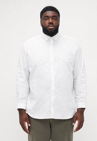 Chemise blanche à manches longues, ornée d'un col, avec une texture lisse et une coupe droite. Les boutons sont de couleur claire, apportant un contraste.
