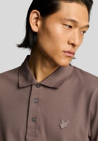 Camisa polo marrón con collar, tres botones, tela texturizada y un pequeño logo bordado en el lado izquierdo del pecho.