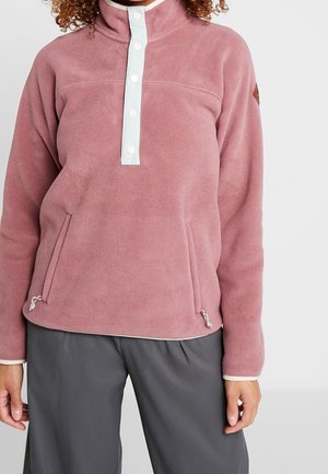 Persoon draagt een stofroze fleece trui met witte drukkers en grijze broek, handen ontspannen langs het lichaam, tegen een effen achtergrond.