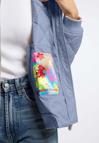 Jasnoniebieska pikowana kurtka z żebrowanymi mankietami, z kolorową kieszenią w kwiatowy wzór. Metka głosi "FRIEDA & FREDDIES NEW YORK."