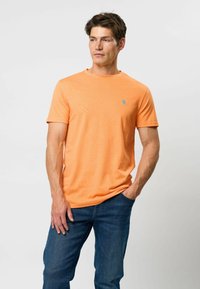 Tricou din bumbac portocalie cu mânecă scurtă, având un mic logo albastru pe piept, asortat cu blugi din denim albastru. Textură netedă și croială lejeră.