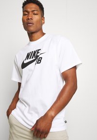 Vit bomullströja med en stor svart Nike SB-logotyp på framsidan. Korta ärmar och standard passform med en etikett på fållen.
