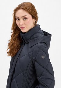 Marineblaues, gesteppte Jacke mit hohem Kragen, strukturierten Schulterakzenten und einem runden Logo-Patch am linken Arm. Glattes, glänzendes Material.