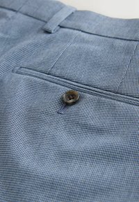 Gros plan sur un pantalon en tissu bleu texturé avec une poche arrière boutonnée et une passants de ceinture sur la ceinture.