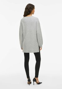 Sieviete ar gariem tumšiem matiem, tērpta brīvā pelēkā džemperī, melnās šaurās biksēs un melnās augstpapēžu kurpēs, skatās prom uz baltā fona.