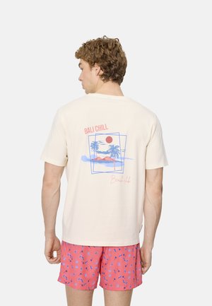Hombre de espaldas que lleva una camiseta color crema con un gráfico de playa que dice "Bali Chill" y pantalones cortos coral con estampado de palmeras azules.