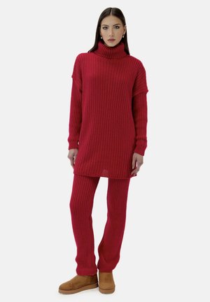 Kvinde iført en rød ribstrikket turtleneck sweaterkjole og matchende leggings med tan ruskind ankelsstøvler, stående mod en hvid baggrund.