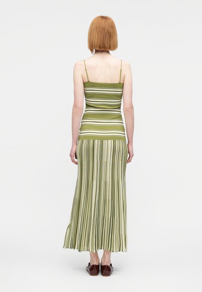 Γυναίκα με ίσια κόκκινα μαλλιά, που φοράει πράσινο και λευκό ριγέ αμάνικο φόρεμα και καφέ παπούτσια, στέκεται με την πλάτη σε απλό φόντο.