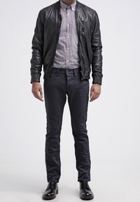 Mann in schwarzer Lederjacke, kariertem Hemd, dunklen Hosen und schwarzen, polierten Schuhen steht vor einem schlichten Hintergrund.