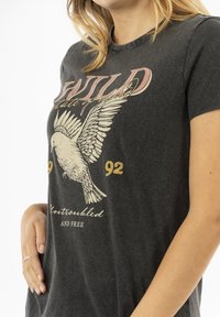 Camiseta gris carbón con cuello redondo, mangas cortas y un gráfico de un pájaro con el texto "WILD" y "92" en acentos de color rosa y oro.