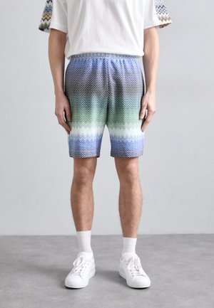 Männer Shorts mit gemustertem Zickzackdesign in Schwarz-, Grün- und Blautönen; elastischer Bund; kombiniert mit weißen Sneakers und Socken.