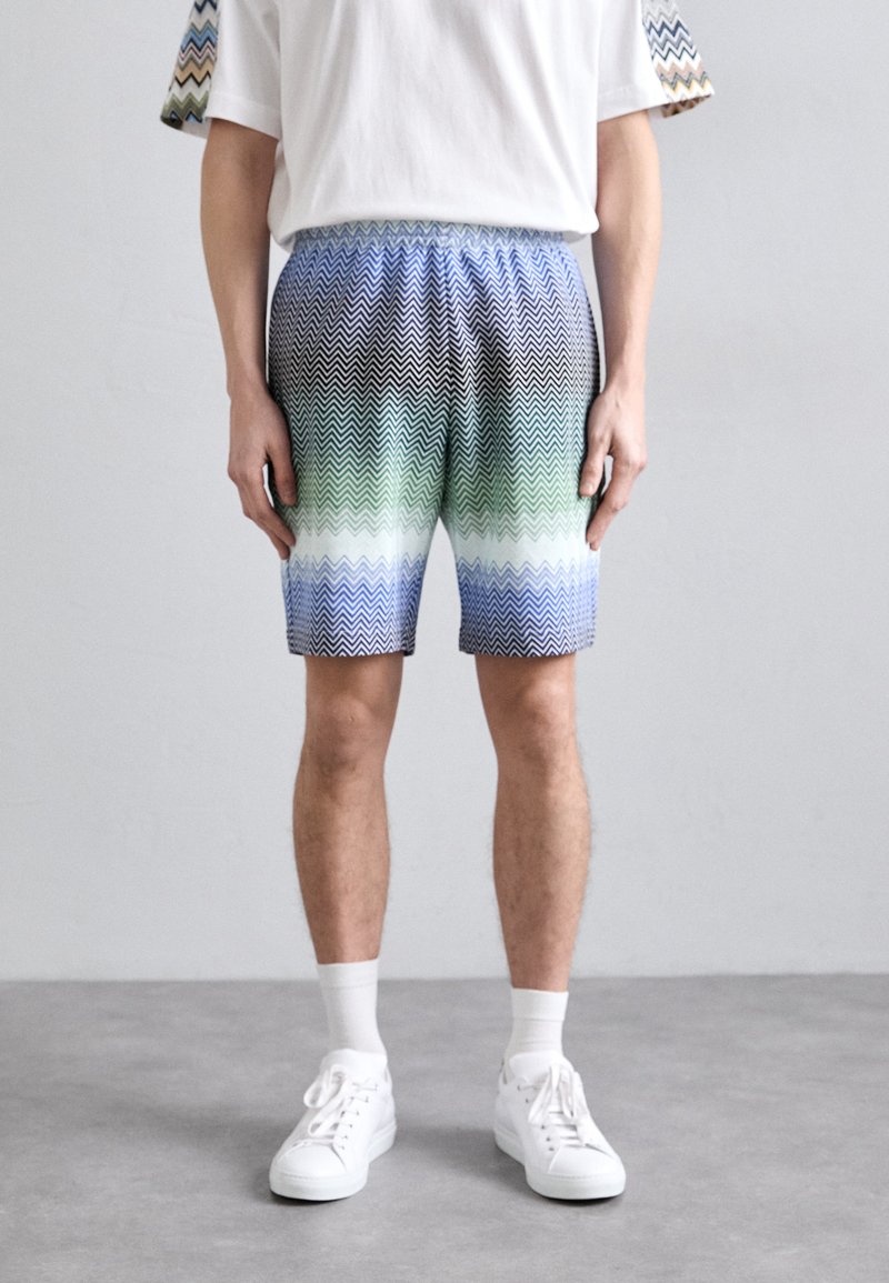 Männer Shorts mit gemustertem Zickzackdesign in Schwarz-, Grün- und Blautönen; elastischer Bund; kombiniert mit weißen Sneakers und Socken.