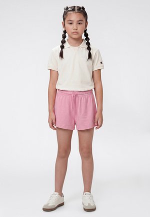 Ein junges Mädchen steht frontal, trägt ein cremefarbenes Poloshirt, pinke Shorts, weiße Turnschuhe und hat geflochtenes Haar vor einem schlichten Hintergrund.