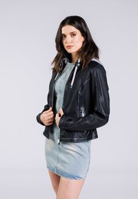 Schwarze Lederjacke mit tailliertem Design. Verfügt über einen Reißverschluss, graue Innenauskleidung und metallische Akzente. Das Model trägt ein helles Denimkleid.