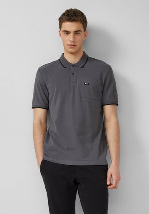 Jeune homme debout contre un mur, portant un polo gris à manches courtes avec une poche poitrine et un pantalon noir, les mains dans les poches.