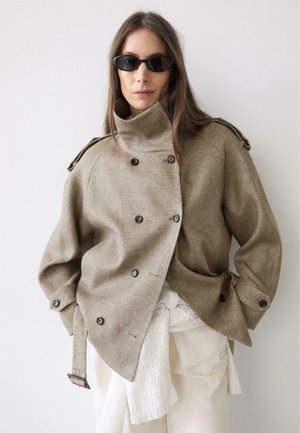 Frau mit langen braunen Haaren trägt übergroßes beige zweireihiges Jackett, schwarze Sonnenbrille und weiße Lagenkleidung vor weißem Hintergrund.