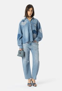 Žena v světle modrém džínsovém oblečení s nášivkami ve tvaru srdce, drží tkanou kabelku a má na sobě tmavě modré sandály na podpatku.