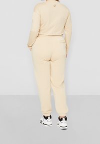 Person in beigen Sweatpants und passendem Langarmoberteil, mit Händen in den Taschen, stehend auf einem grauen Boden vor einer weißen Wand.