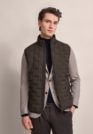 Padded vest in dark olive green with a high collar, zip front, and quilted texture, layered over a light gray suit jacket.