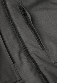 Grå tyg med en slät textur som har två svarta dragkedjor. Dragkedjorna är placerade vid sidan av fickorna, vilket betonar funktionalitet och design.