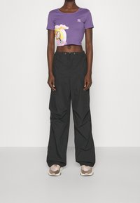 Fioletowy skrócony t-shirt z graficznym nadrukiem przedstawiającym tęczę i postać. Czarna, luźna, cargo pants. Neutralne trampki.