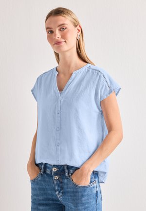 Jeune femme aux cheveux blonds porte un chemisier bleu clair à manches courtes boutonné et un jean bleu, les mains dans les poches, sur un fond uni.