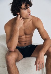 Boxeri din bumbac negri, cu un elastic alb cu logo-ul Calvin Klein, purtați de un model masculin așezat, cu păr creț și o fizionomie tonifiată.