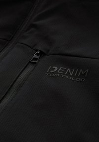 Krupan kadar crne tkanine jakne s patentnim zatvaračem i reljefnim logotipom "DENIM TOM TAILOR" na području prsiju.