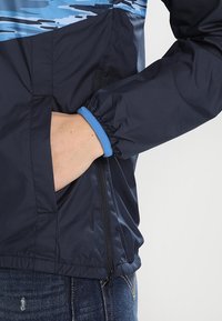 Personne portant une veste bleu marine avec un poignet bleu, main dans la poche avant, et un jean en denim bleu.