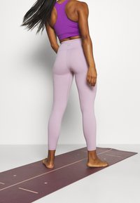 Světlé fialové legíny s vysokým pasem a hladkým povrchem, kombinované s přiléhavým tmavě fialovým tílkem, stojící na podložce na jógu.