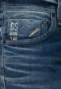 Siniset farkut, joissa on kulunut etutasku, jossa on valkoinen brodeeraus tekstillä "CS RAW 3 101" ja beige ommelkoriste.