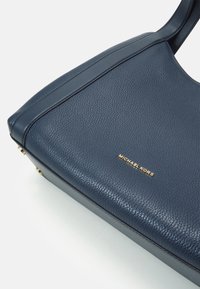 Mörkblå texturerad läderhandväska med guld "Michael Kors"-logotyp och guldfärgade metallfötter i nederkant.