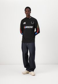 Svart langermet skjorte med "NASCAR"-logo og fargerike striper på ermene, parret med mørk denim jeans og beige sneakers.