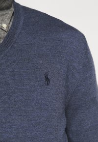 Pull col en V bleu marine en tissu texturé, avec un logo subtil sur la poitrine, porté sur une chemise grise à col.