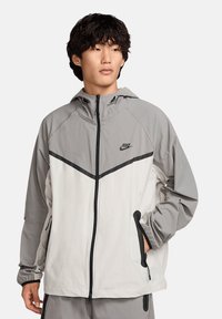 Nike jasje met een grijs-witte kleurencombinatie, lichtgewicht stof, ritssluiting, verstelbare kap en contrasterende accenten langs de naden.