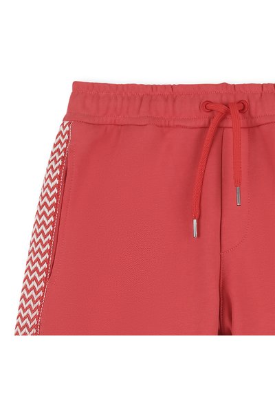 Shorts avec taille élastique rouge, cordon de serrage et poches latérales, présentant un motif en zigzag rouge et blanc sur les panneaux latéraux.