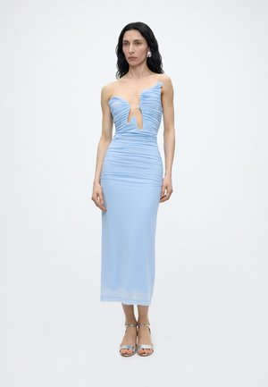 Γυναίκα με μακριά σκούρα μαλλιά, φορώντας ένα ανοιχτό μπλε μίντι φόρεμα χωρίς τιράντες με ρυτίδες και ασημένια ανοιχτά παπούτσια, στέκεται μπροστά σε απλό λευκό φόντο.