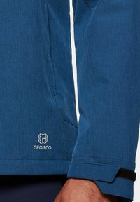 Niebieska kurtka z tkaniny o teksturowanym wyglądzie, z białym logo Geo Eco w pobliżu dołu oraz regulowanymi mankietami z czarnym paskiem na rzep.