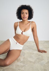 Two-piece bikini in off-white textured fabric. Top features a triangular shape and contrasting brown hardware. High-waisted bottoms.