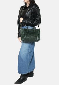Borsa in pelle verde con motivo coccodrillo, con doppi manici, forma rettangolare e tracolla regolabile. Indossata da una persona in abbigliamento nero e blu.