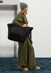Fekete válltáska cipzáras zsebbel, puha, texturált anyagból készült. A modell olívazöld ruhát és szürke kötött sapkát visel.
