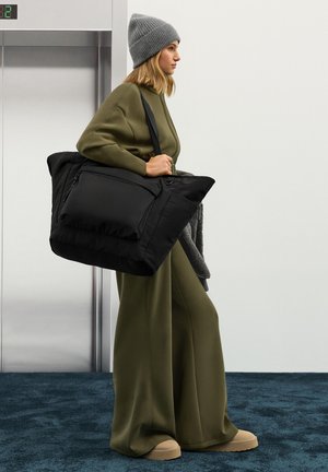 Schwarze Tragetasche mit Reißverschlusstasche, hergestellt aus weichem, strukturiertem Stoff. Das Modell trägt ein olivgrünes Outfit und eine graue Strickmütze.