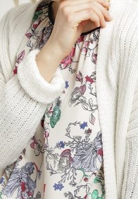 Vit stickad cardigan med texturerat mönster, lagd över en blommig topp med rosa, gröna och blå accenter på en krämfärgad bakgrund.