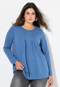 Blaues langärmeliges Oberteil aus weichem Stoff, mit Falten und subtilen Details am Ausschnitt, kombiniert mit grauen Jeans.