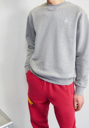 Osoba ubrana w szarą bluzę z białym logo Jumpman oraz czerwone spodnie dresowe z małym żółtym wzorem na udzie.
